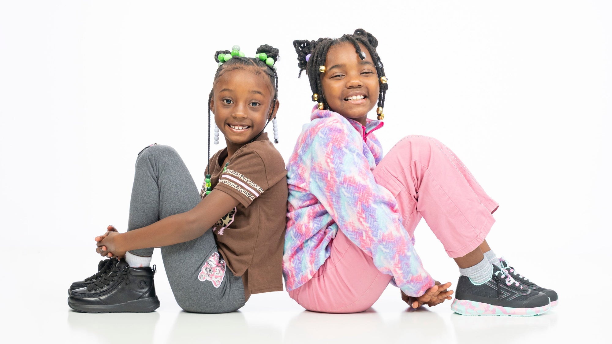 Two young girls sitting back-to-back on a white studio floor, wearing casual streetwear. The girl on the left wears a brown t-shirt, gray leggings, and black sneakers.