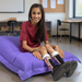 A smiling young woman sits on a large purple bean bag chair in a classroom setting. She is wearing a maroon t-shirt, black socks with a floral pattern, and black sneakers.