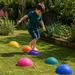 A young boy in a green shirt and navy shorts balances barefoot on a colorful dome-shaped exercise ball in a sunny backyard garden, with other matching balls on the grass.